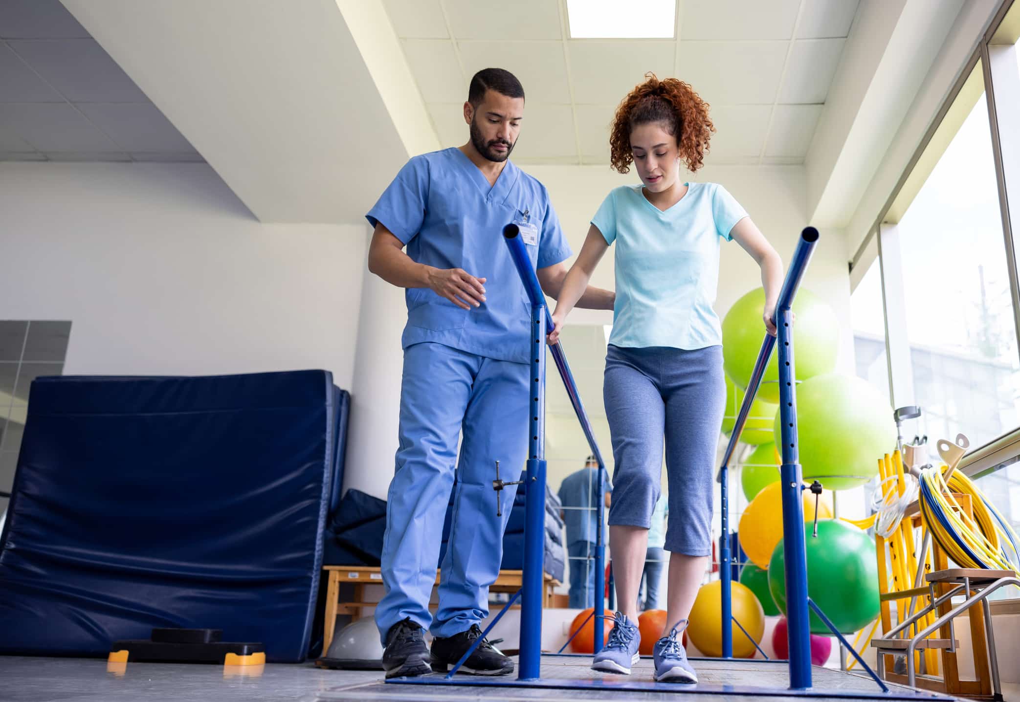 Physiotherapist assisting patient walking between parallel bars in rehabilitation clinic